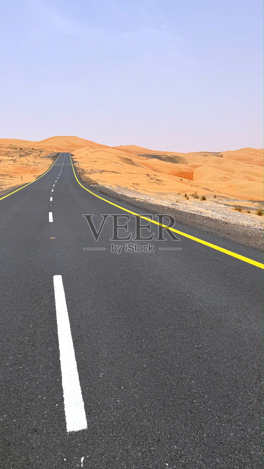 长长的沥青公路穿越深邃的沙漠，路面有白色和黄色的条纹，背景是沙丘，阳光明媚，天空晴朗。照片摄影图片