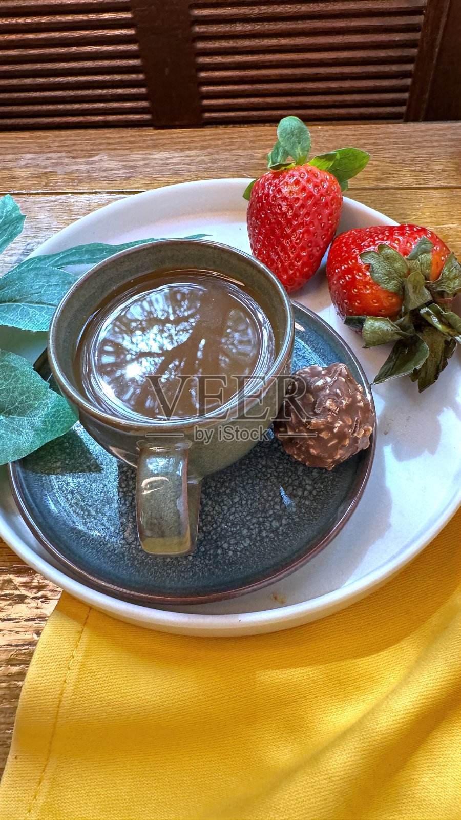 蒸汽腾腾的咖啡杯静静地放在碟子上，旁边是成熟的草莓和一块黑巧克力，展现出温馨的早餐时光，背景是磨损的木质表面和柔软的黄色餐巾。照片摄影图片
