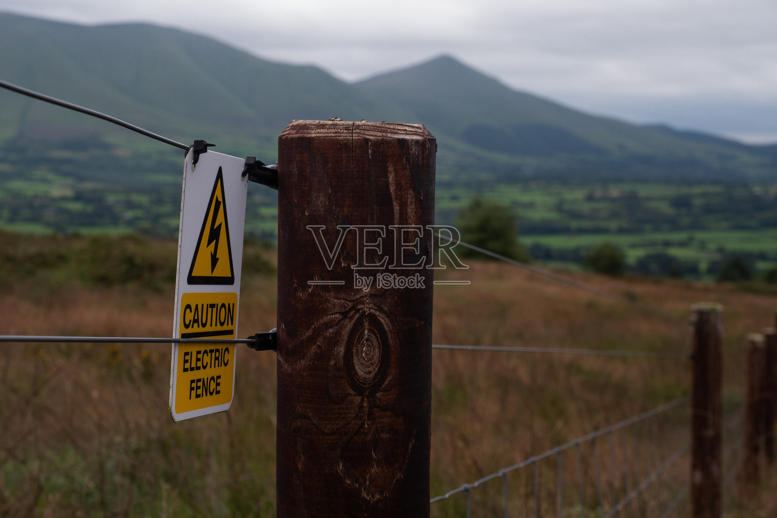 警告标志贴在木柱上，标示着电围栏，背景是起伏的山丘和多云的乡村天空。照片摄影图片