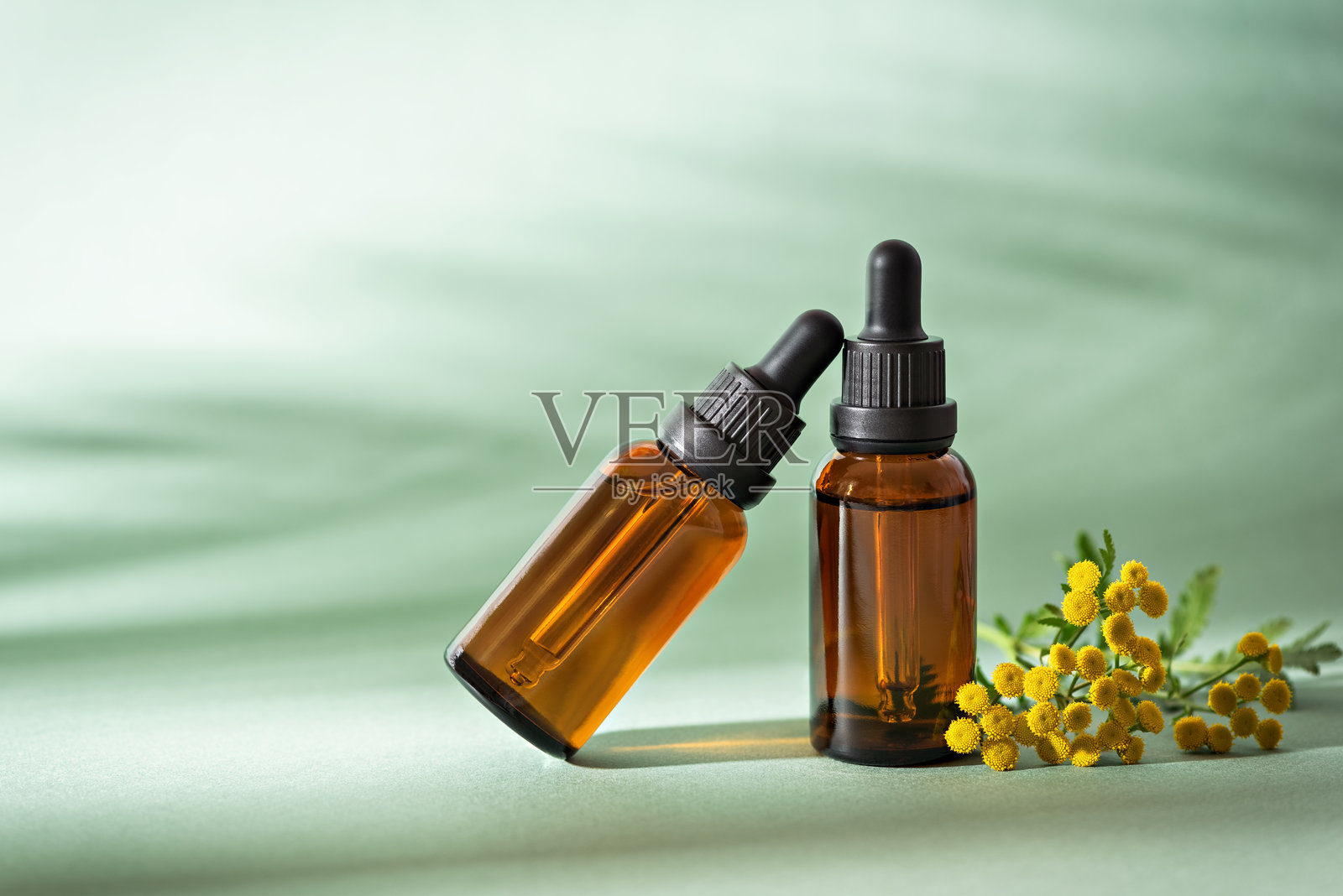 带有黄色千屈菜花装饰的琥珀色滴管瓶，瓶内含有精华液或精油，背景为绿色，带有阴影，化妆品护肤产品照片摄影图片