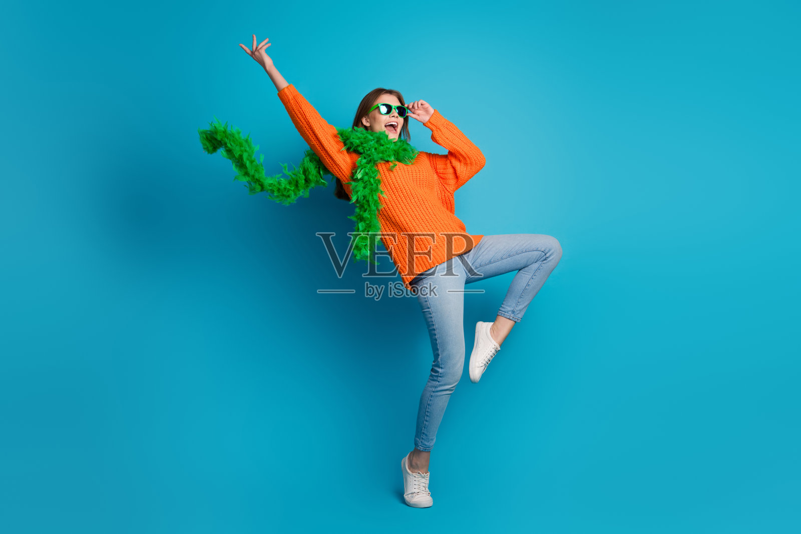 穿着鲜艳橙色毛衣的快乐年轻女性在明亮的蓝色背景前摆出俏皮的姿势，展现时尚风格照片摄影图片