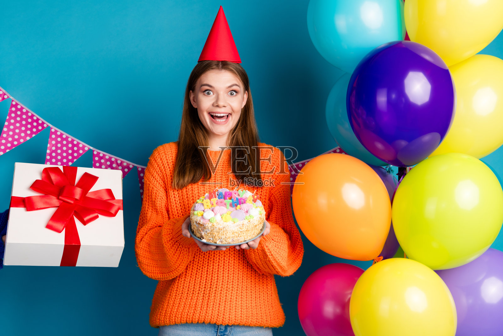 美丽的年轻女性在充满活力的蓝色墙壁前庆祝生日，周围有五彩缤纷的气球、礼物和蛋糕。照片摄影图片