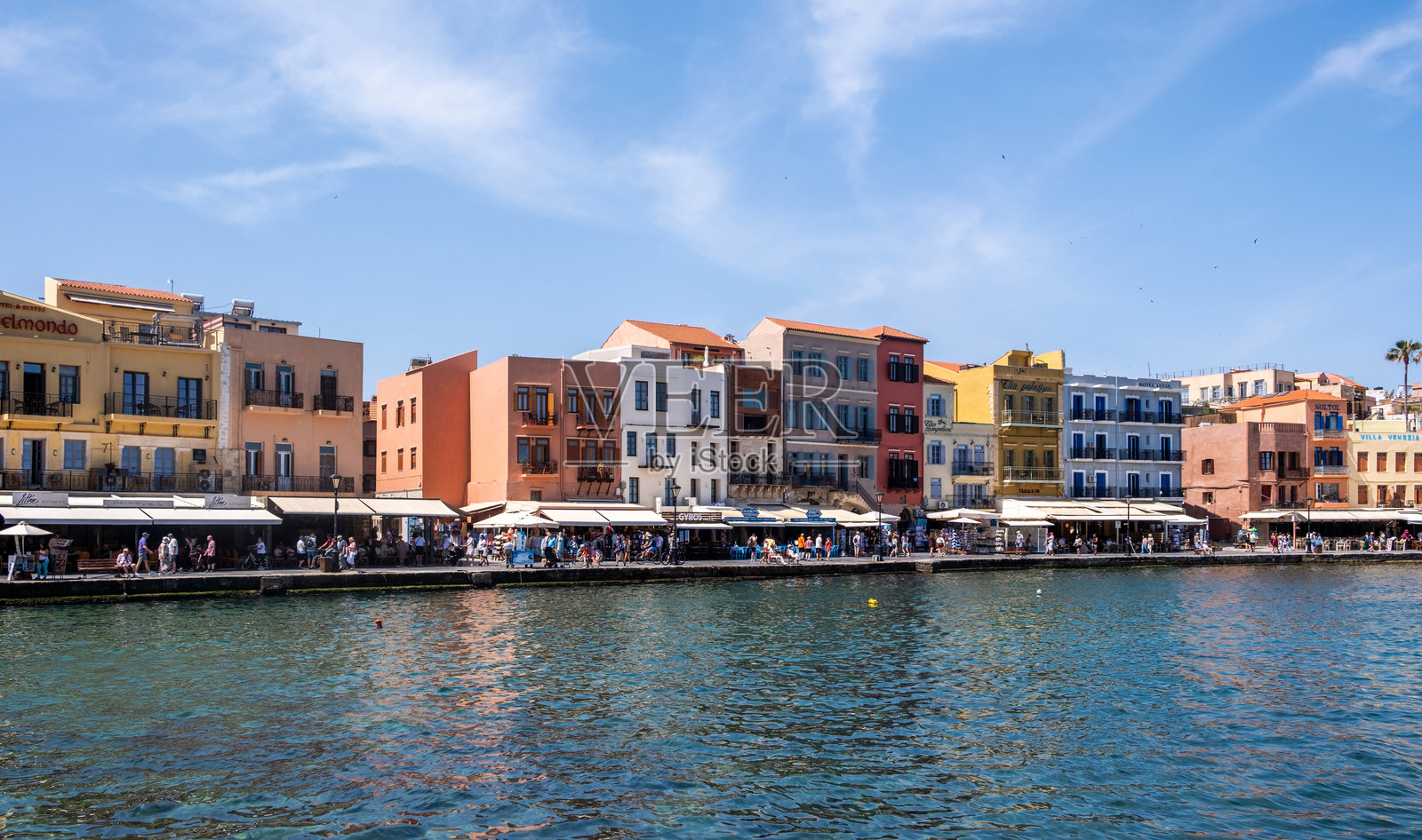 希腊哈尼亚。威尼斯风格的老港口建筑位于海滨长廊，行人漫步在咖啡馆和商店旁，蓝天。照片摄影图片