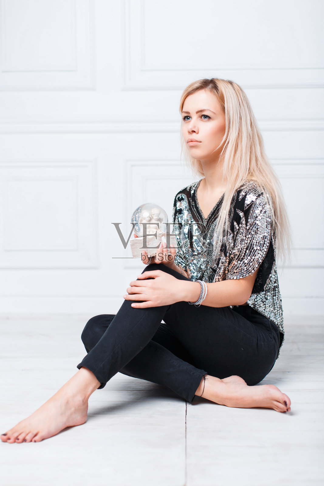 年轻漂亮的金发女孩坐在地板上，手中拿着一个有天使的魔法圣诞球。照片摄影图片