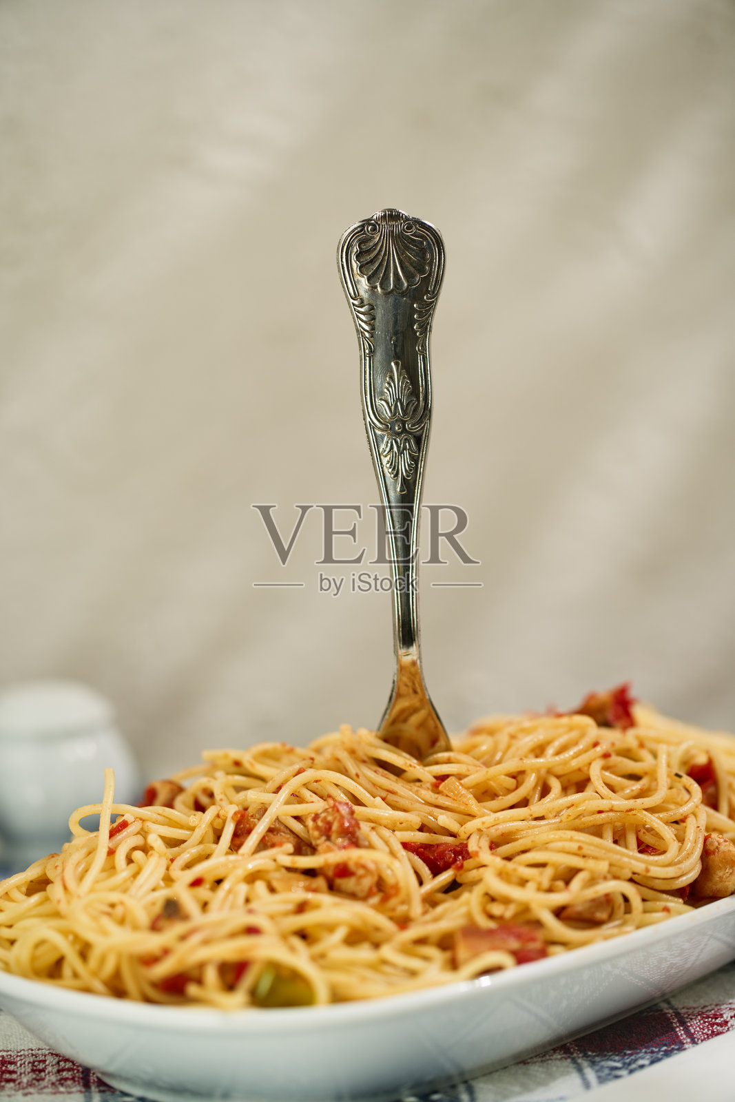 在一面被藤叶阴影点缀的石灰洗墙前，一把叉子直立在满是意大利面的盘子里，桌子上铺着地中海图案的布。照片摄影图片