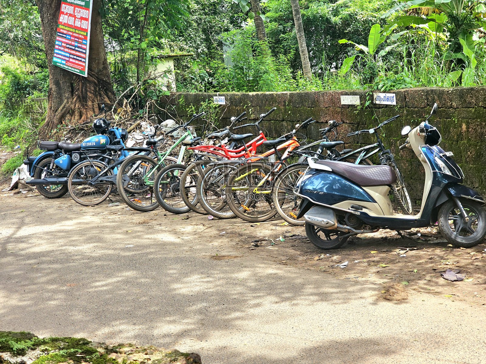 户外环境中，几辆自行车、摩托车和一辆滑板车停靠在石墙和郁郁葱葱的树木旁。照片摄影图片