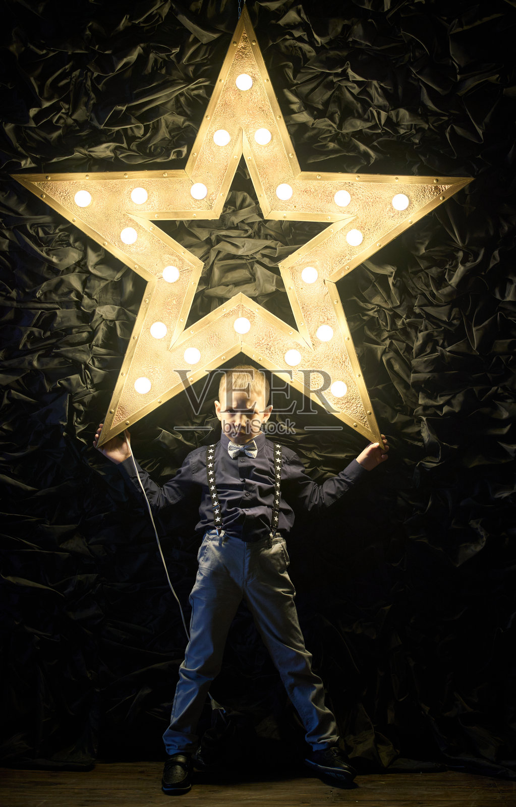 一个穿着正式服装、系着领结的严肃男孩，背靠在灯下的墙上。照片摄影图片