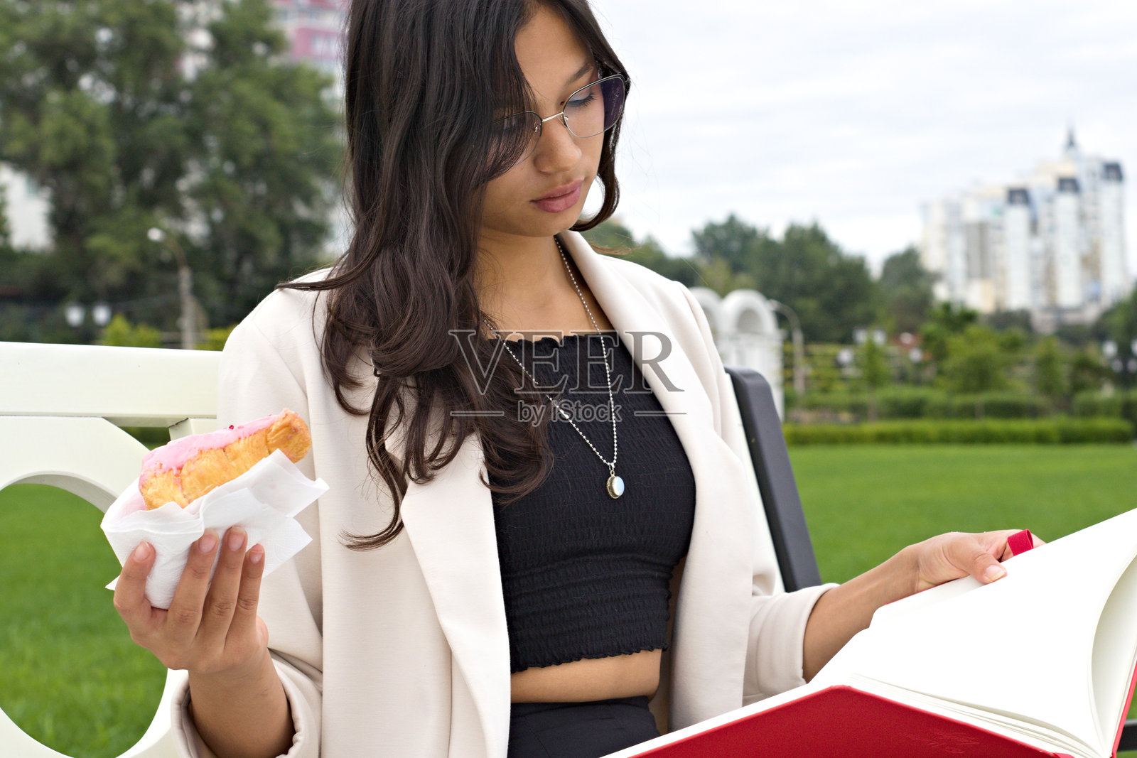 年轻的印度大学生坐在户外长椅上，阅读一本书，吃着可颂，象征着现代学术生活方式，学习、休闲与追求知识的平衡。照片摄影图片