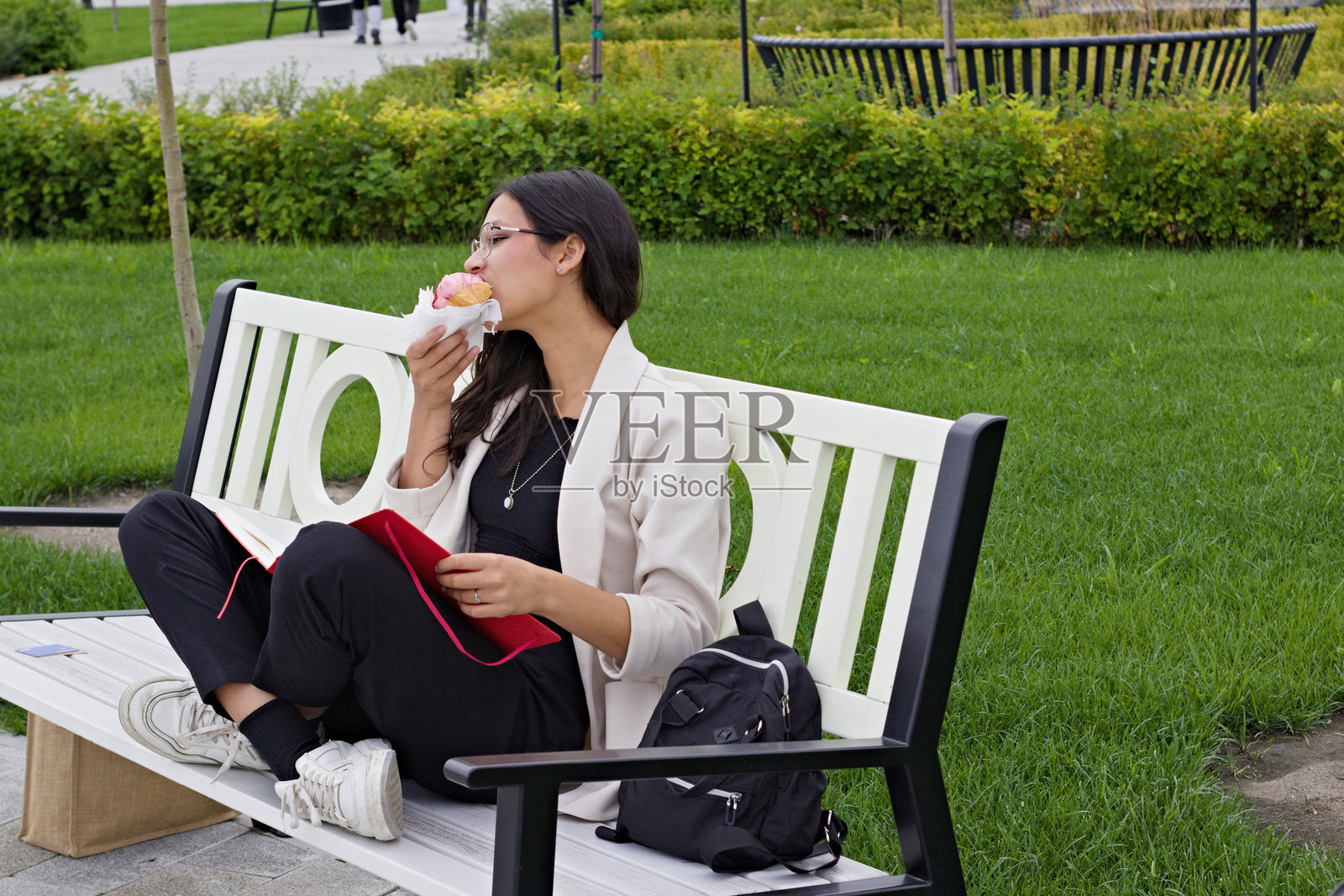 年轻的印度大学生坐在户外长椅上，阅读一本书，吃着可颂，象征着现代学术生活方式，学习、休闲与追求知识的平衡。照片摄影图片