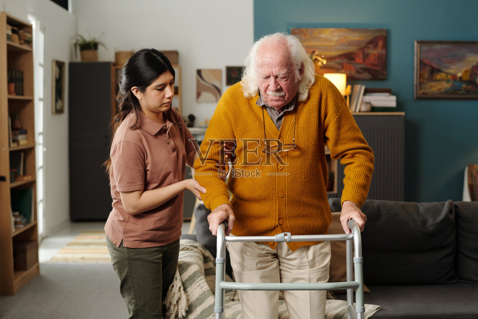 年轻的西班牙裔女性在室内帮助一位年长的白人男性使用助行器照片摄影图片