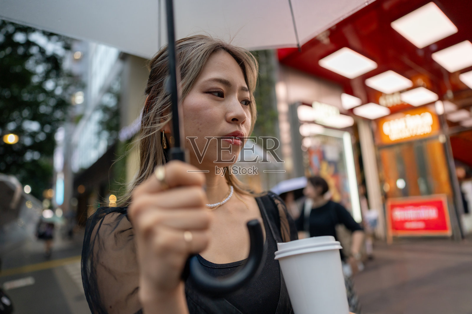 年轻女子穿着黑色连衣裙在雨天的城市中行走，手里拿着咖啡杯照片摄影图片