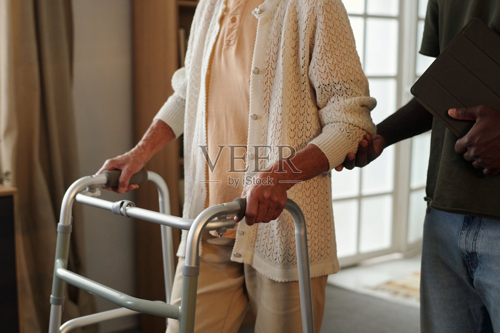 一位年长的白人女性在室内使用助行器，由一位年轻的黑人男性协助。照片摄影图片