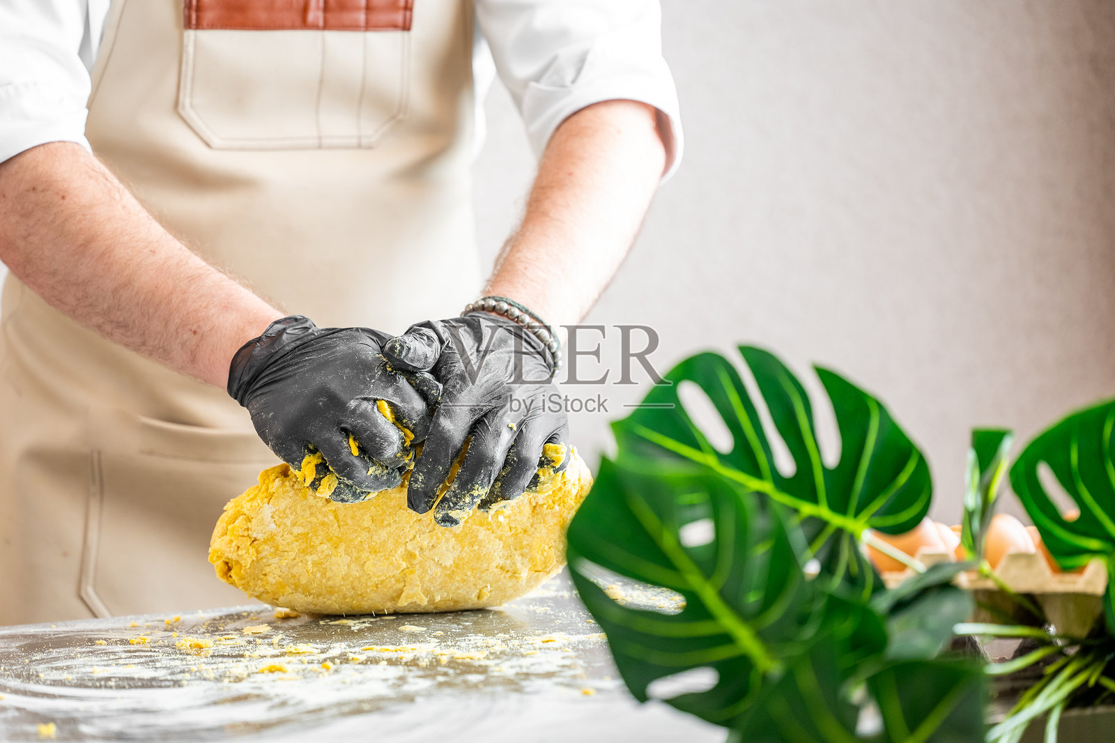 厨师在撒有面粉的台面上揉黄色面团，穿着手套和围裙。新鲜的食材和龟背竹植物在前景中。乡村厨房环境中的烹饪准备。照片摄影图片