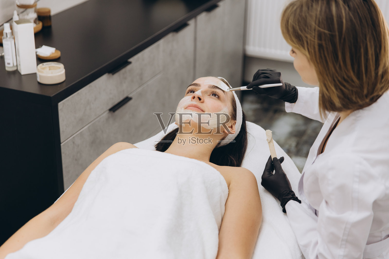 美容师为躺在按摩床上的年轻女性涂抹面膜照片摄影图片