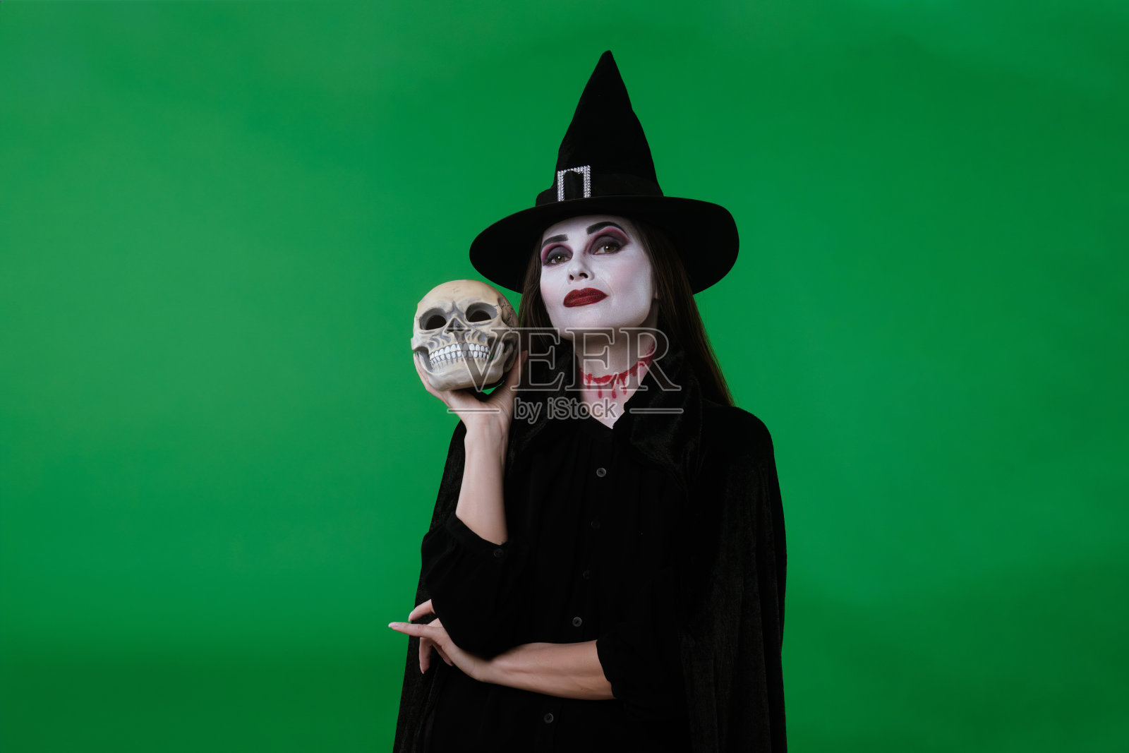 一位穿着黑色斗篷和帽子的吸血鬼女巫在绿色背景的摄影棚中与骷髅摆姿势。照片摄影图片