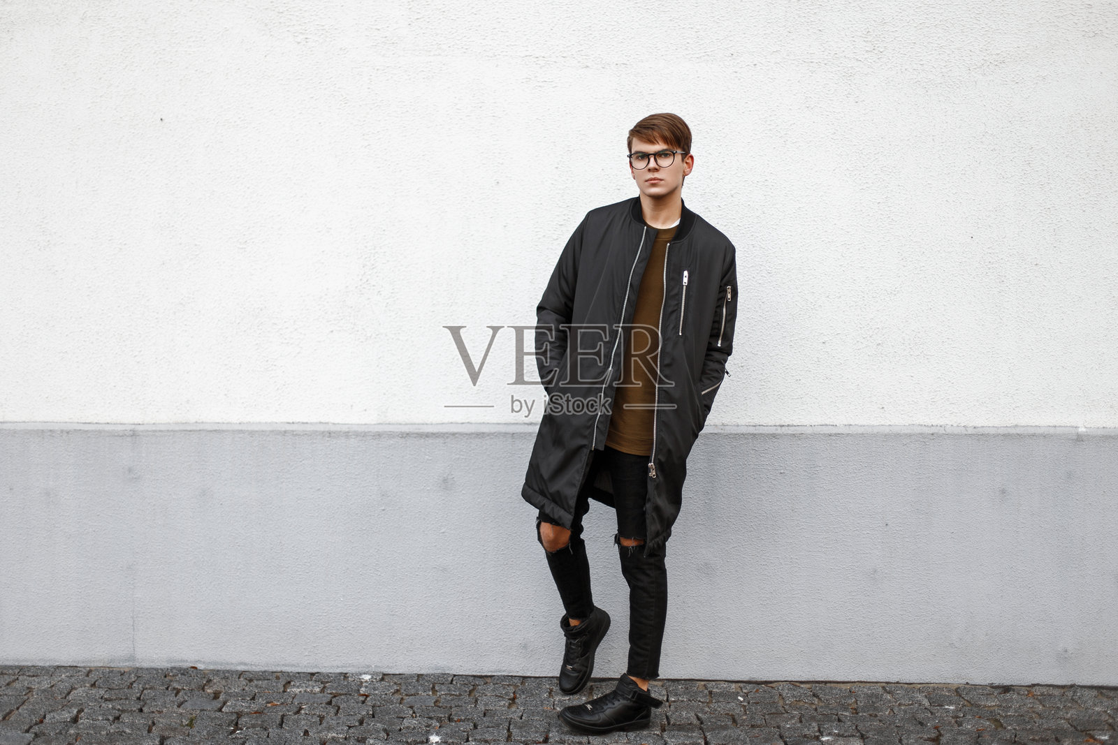 年轻模特男孩戴着眼镜，穿着黑色夹克和破洞牛仔裤，站在墙边。照片摄影图片