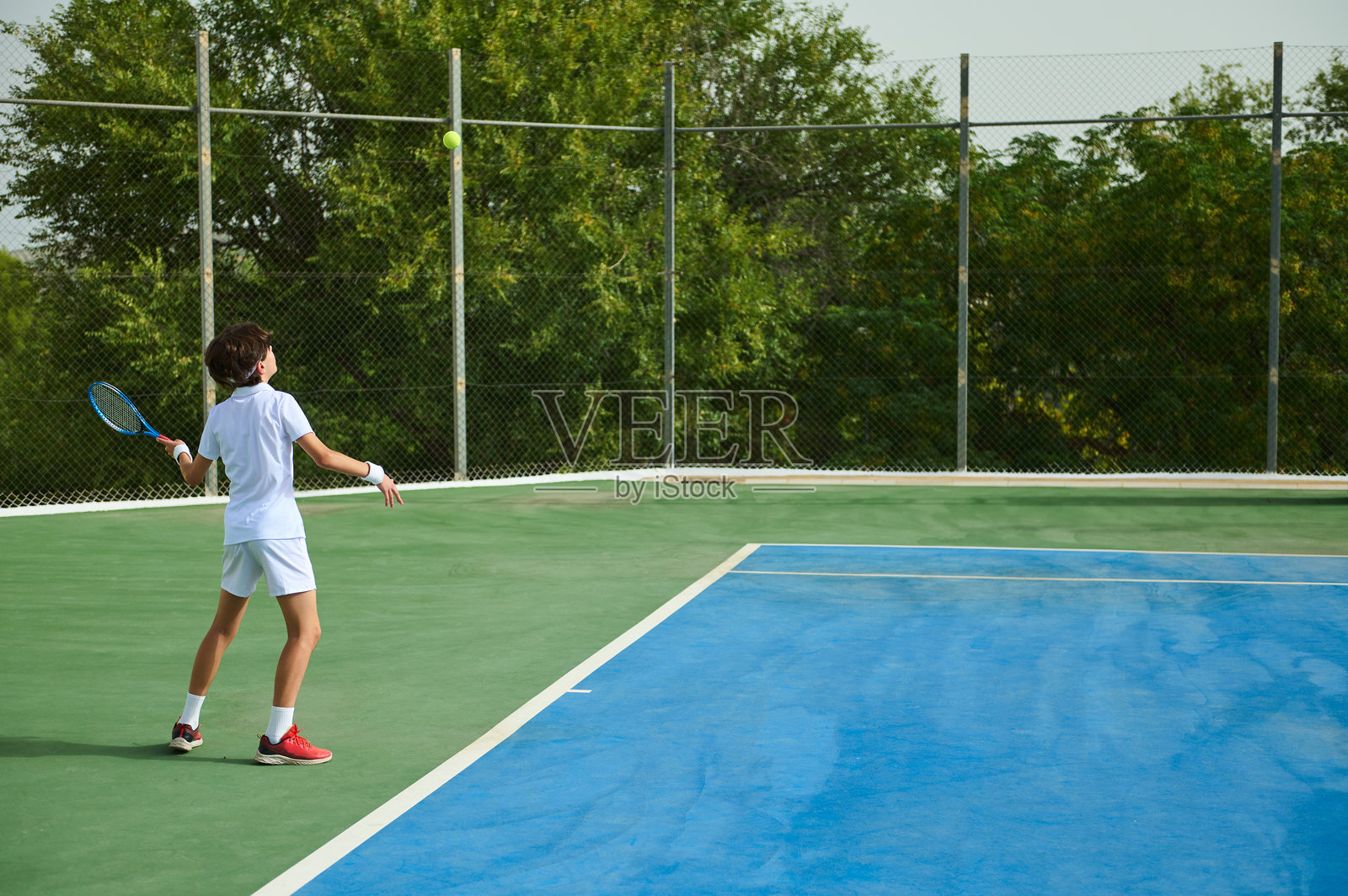 年轻的网球运动员在户外球场发球，周围被绿树环绕照片摄影图片
