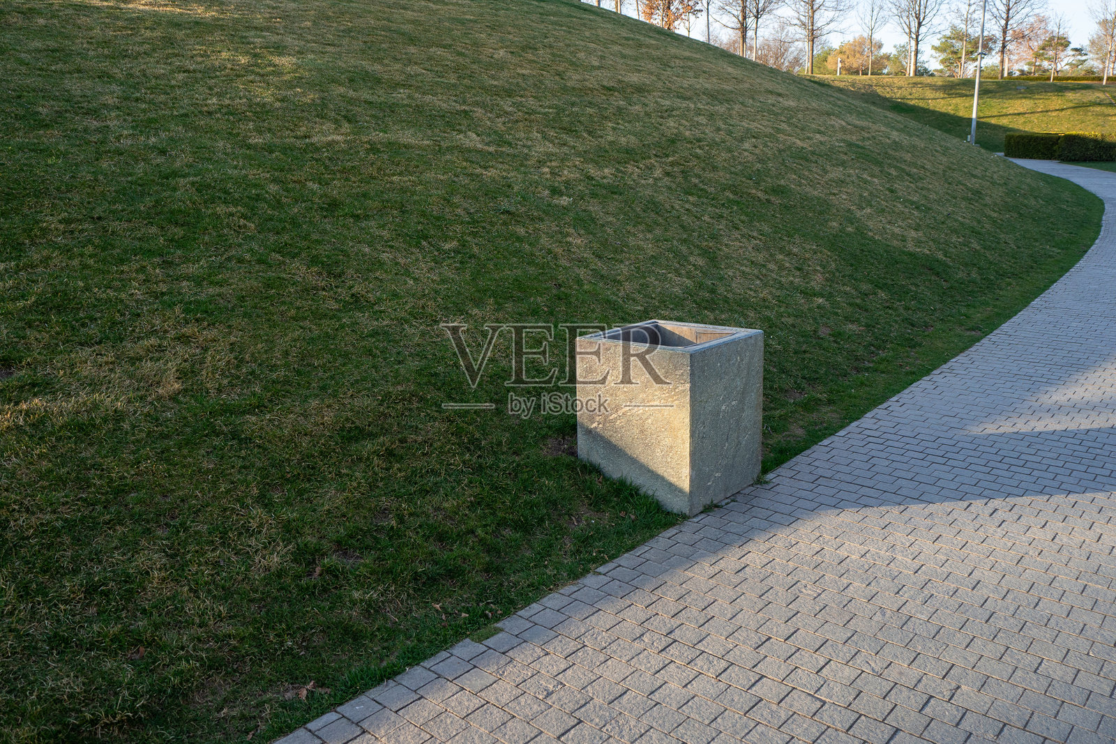 公园环境中，铺砌小径旁放置的混凝土垃圾桶，周围环绕着草地。下午的阳光照耀，突显城市景观元素。照片摄影图片