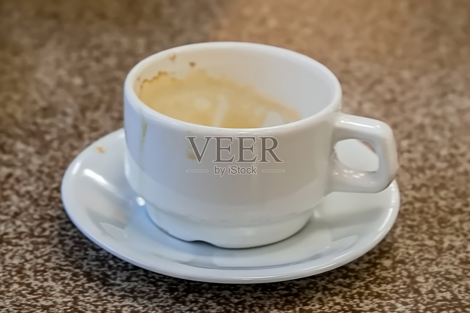 一个使用过的白色浓缩咖啡杯放在一个配套的碟子上，杯中残留着咖啡，背景是一个带有斑点的桌面。照片摄影图片