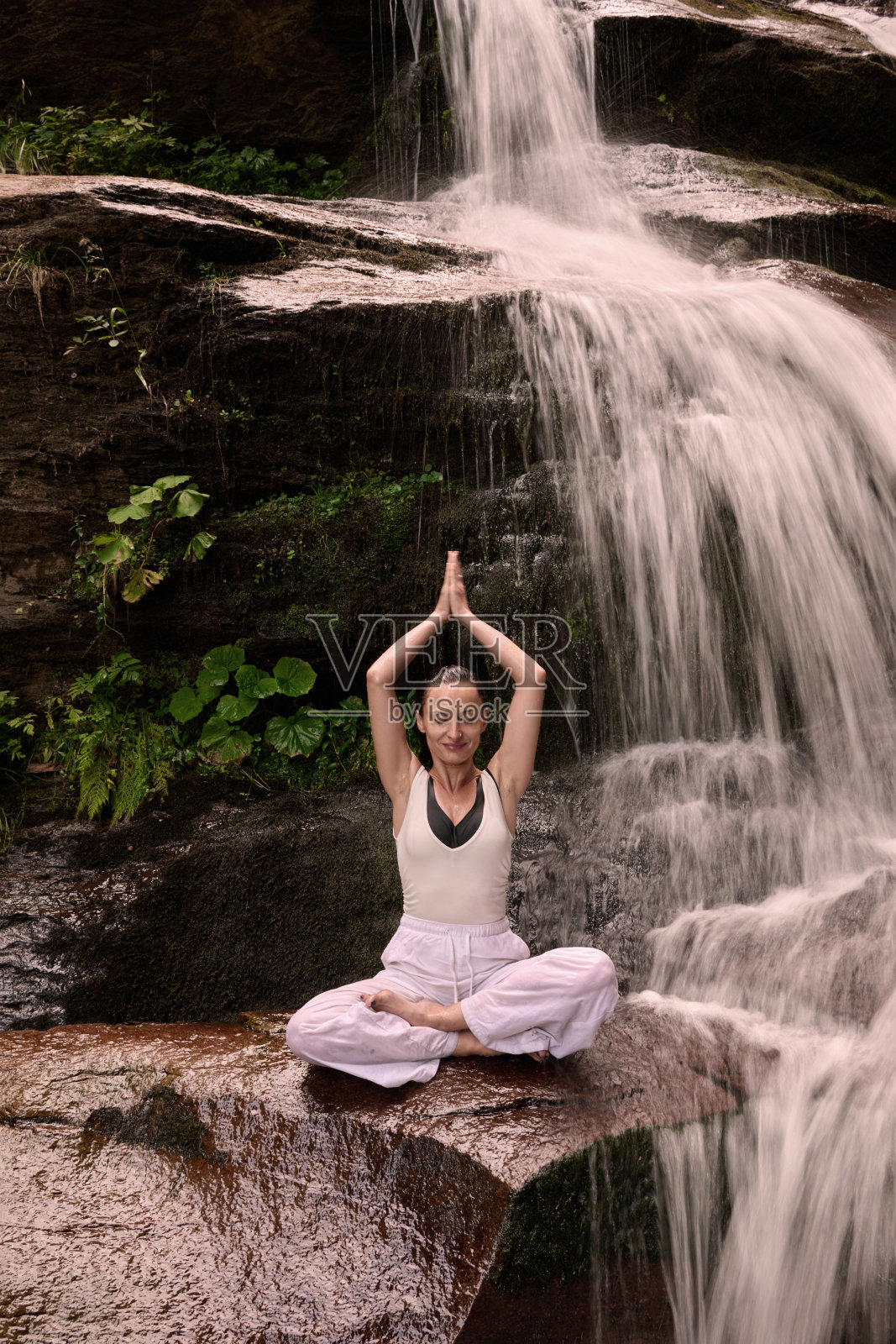 年轻女性坐在热带瀑布下的莲花坐姿中，冥想时展现出平静的力量和正念，完全与自然的能量、宁静和内心的平衡相连接。照片摄影图片
