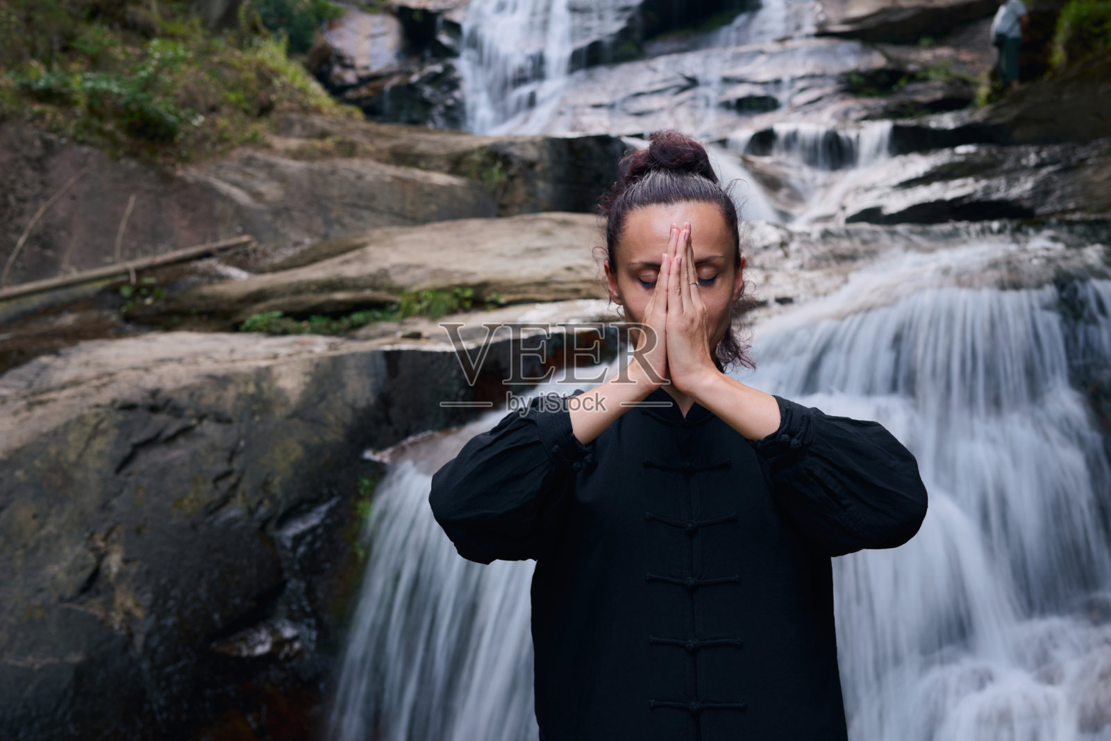 Woman practicing Tai Chi in nature, wearing a traditional black martial arts uniform, standing gracefully near a waterfall, focused on meditation and balance. Healthy lifestyle in asian culture照片摄影图片