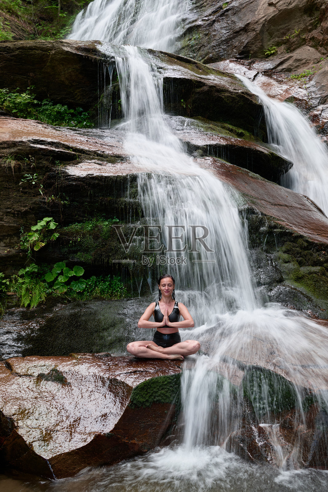 年轻女性坐在热带瀑布下的莲花坐姿中，冥想时展现出平静的力量和正念，完全与自然的能量、宁静和内心的平衡相连接。照片摄影图片