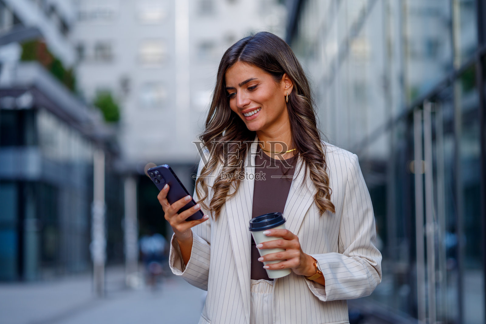 微笑的女商人在城市中走动，一边享用咖啡一边使用智能手机，散发出自信与时尚。照片摄影图片