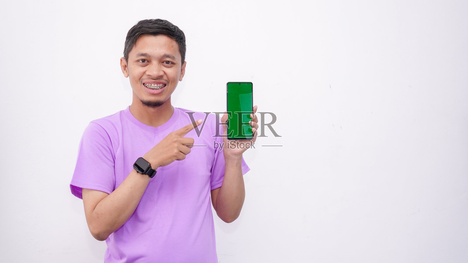 亚洲男子手持智能手机，向镜头展示绿色屏幕并指向你，快乐的千禧一代男孩推荐新应用程序或移动网站，样板横幅拼贴照片摄影图片