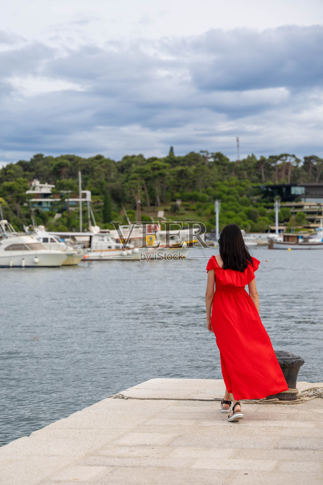 穿着红色连衣裙的女人在码头旁散步。一位穿着飘逸红裙的女人沿着码头走，面朝着布满船只的码头，天空阴云密布。照片摄影图片
