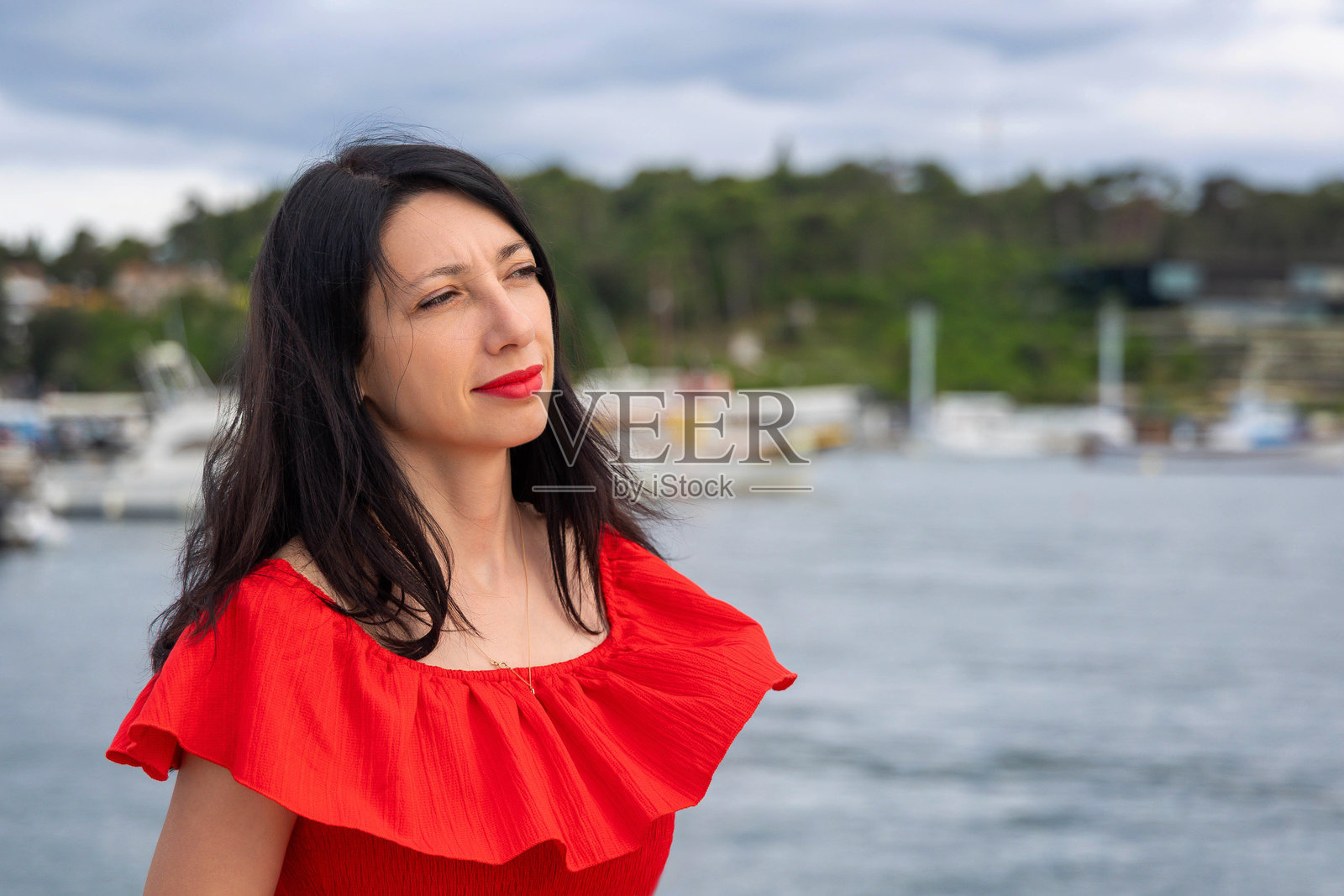 身穿红色连衣裙的沉思女性在水边。她站在海边，凝视着远方，背景中有船只。照片摄影图片