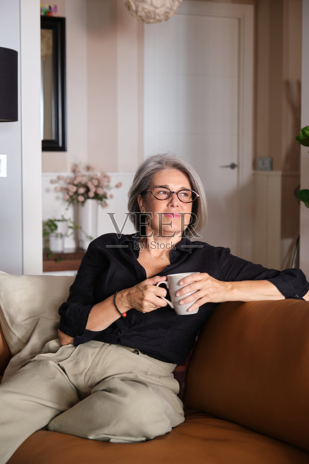 一位年长女性坐在沙发上放松，手中拿着一杯咖啡或茶，目光投向远方。照片摄影图片