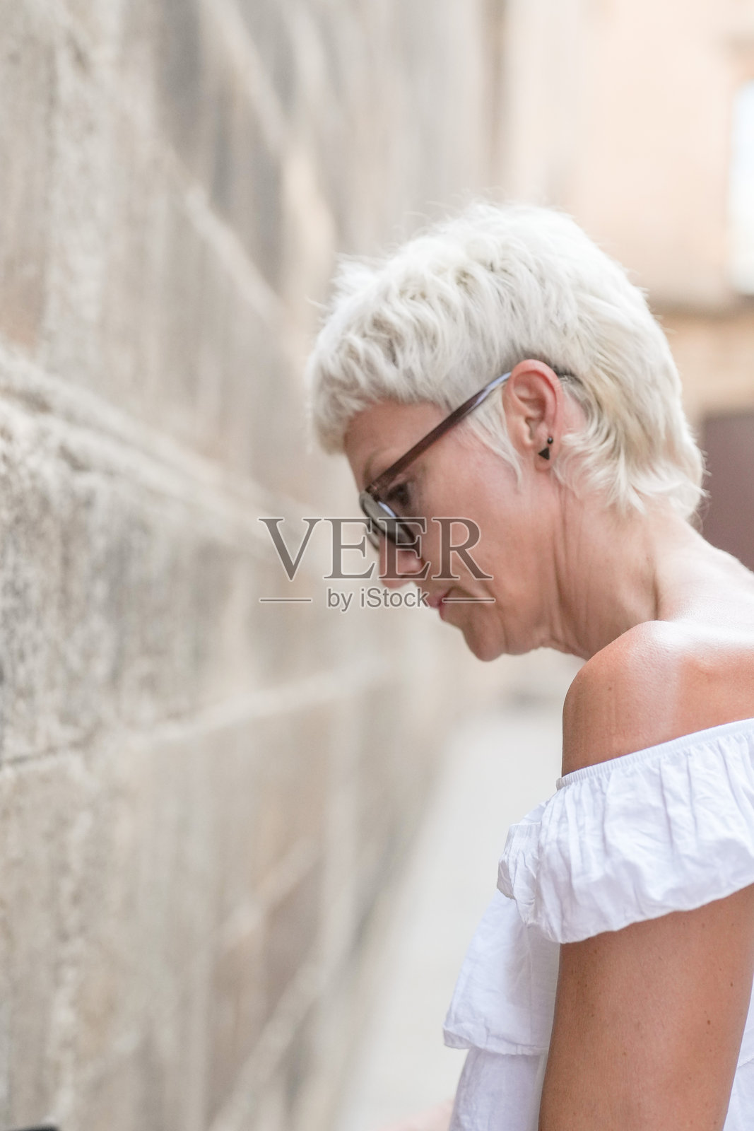 一位短发金发、戴眼镜的女性站在砖墙前。照片摄影图片