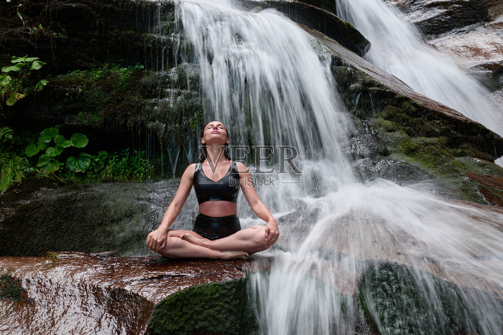 年轻女性坐在热带瀑布下的莲花坐姿中，冥想时展现出平静的力量和正念，完全与自然的能量、宁静和内心的平衡相连接。照片摄影图片
