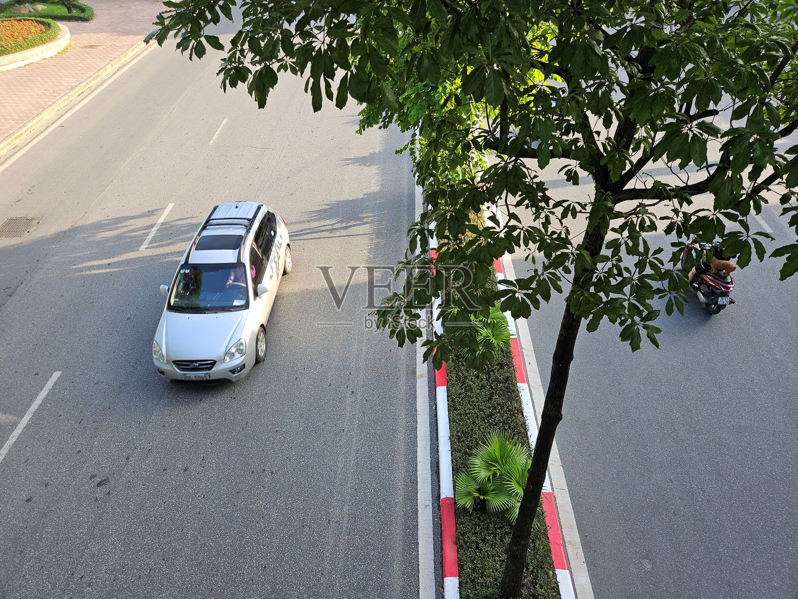 高处俯瞰一条宽阔的城市道路,白色汽车和摩托车驶过一棵绿树和中央隔离带。照片摄影图片