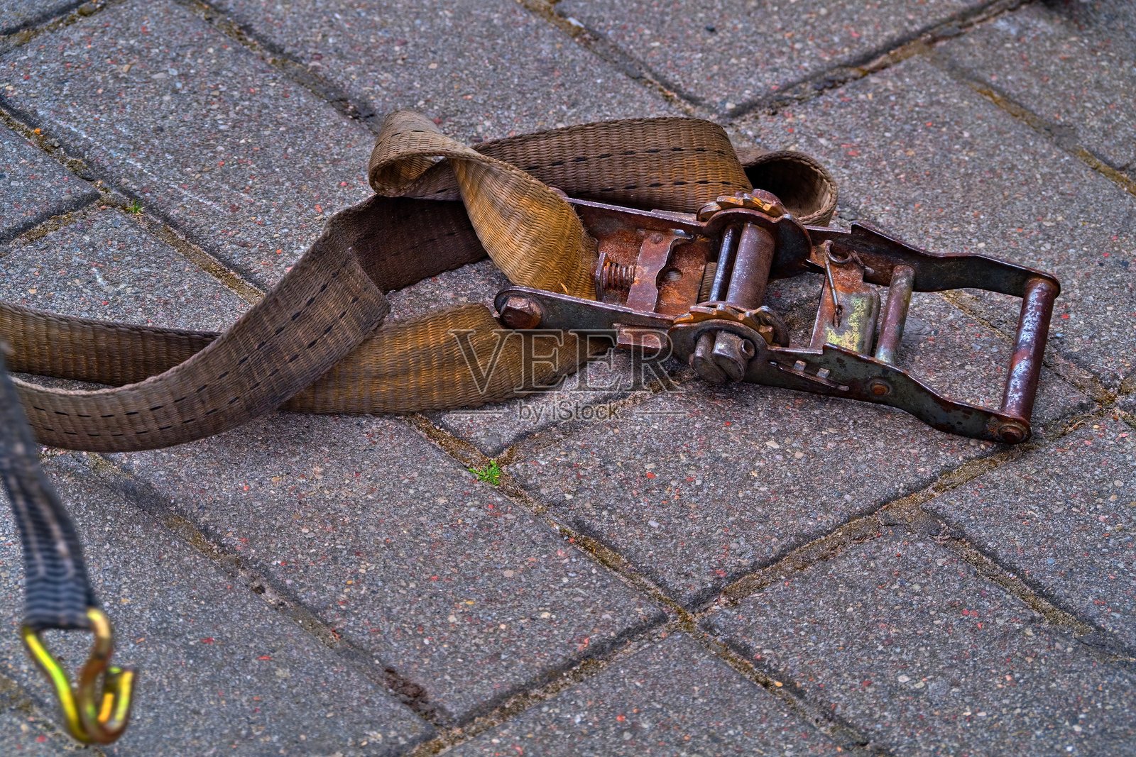 一条旧的生锈的棘轮带特写，旁边是一条磨损的皮带，躺在石铺路面上。这是用于运输和物流的货物固定设备。照片摄影图片