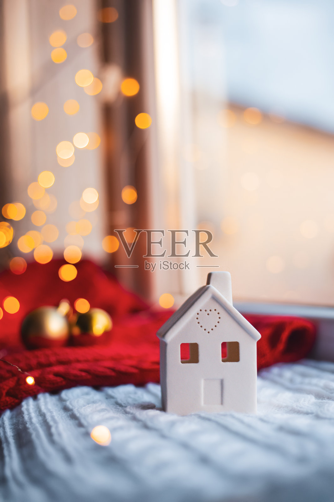 白色陶瓷房子，心形设计，放在温暖的针织毛毯上，窗边有金色圣诞灯光和红色节日背景，象征着温馨的家和节日氛围。照片摄影图片