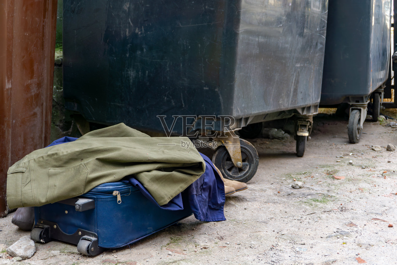 被遗弃的行李箱和衣物放在大型塑料垃圾箱旁，受损的院子地面，被忽视的城市环境，贫困的象征。照片摄影图片