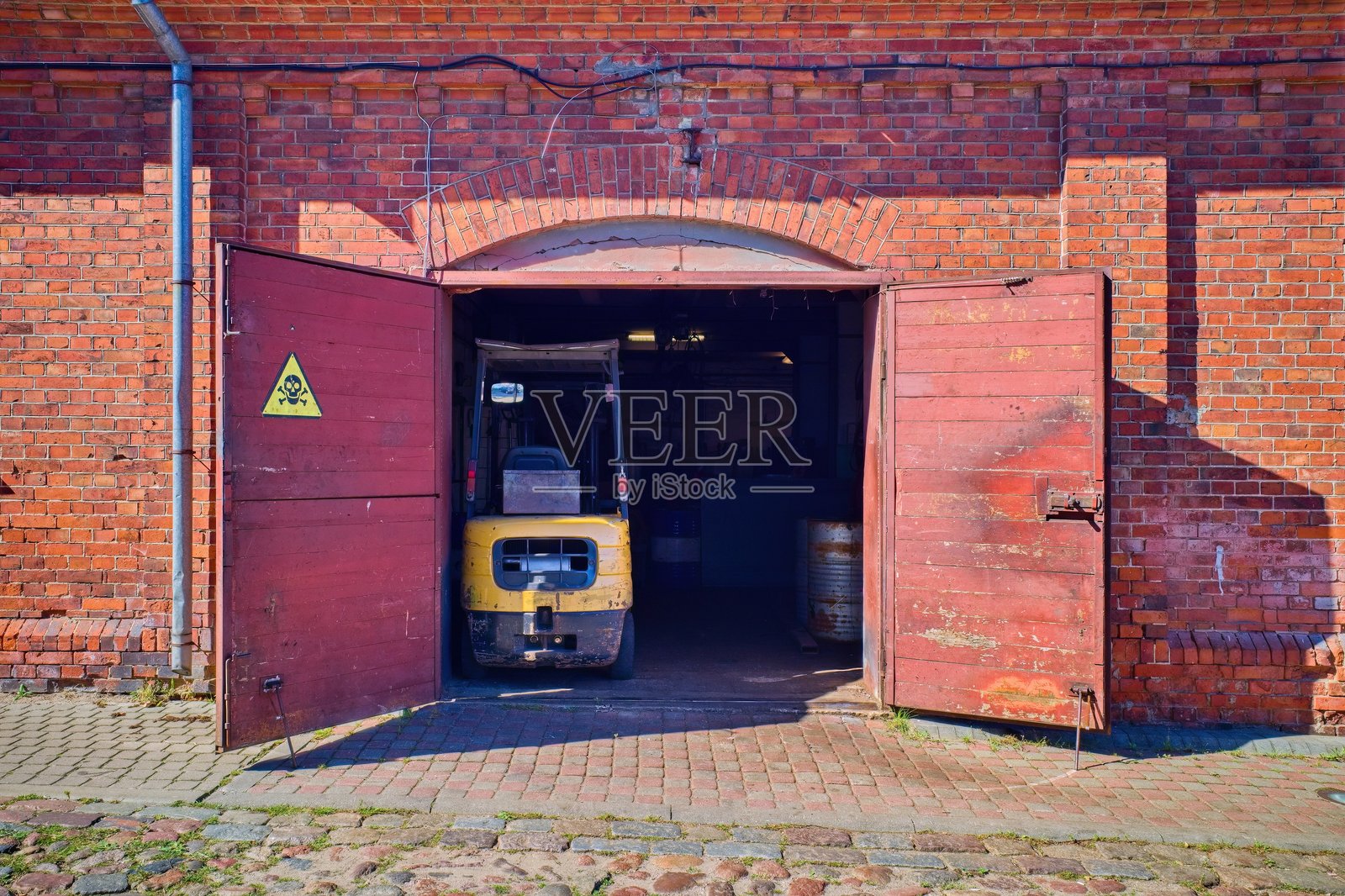 老旧的红砖工业建筑，仓库内有敞开的木门和一辆黄色的叉车。门上可见一个带有骷髅和交叉骨头的警告标志。照片摄影图片