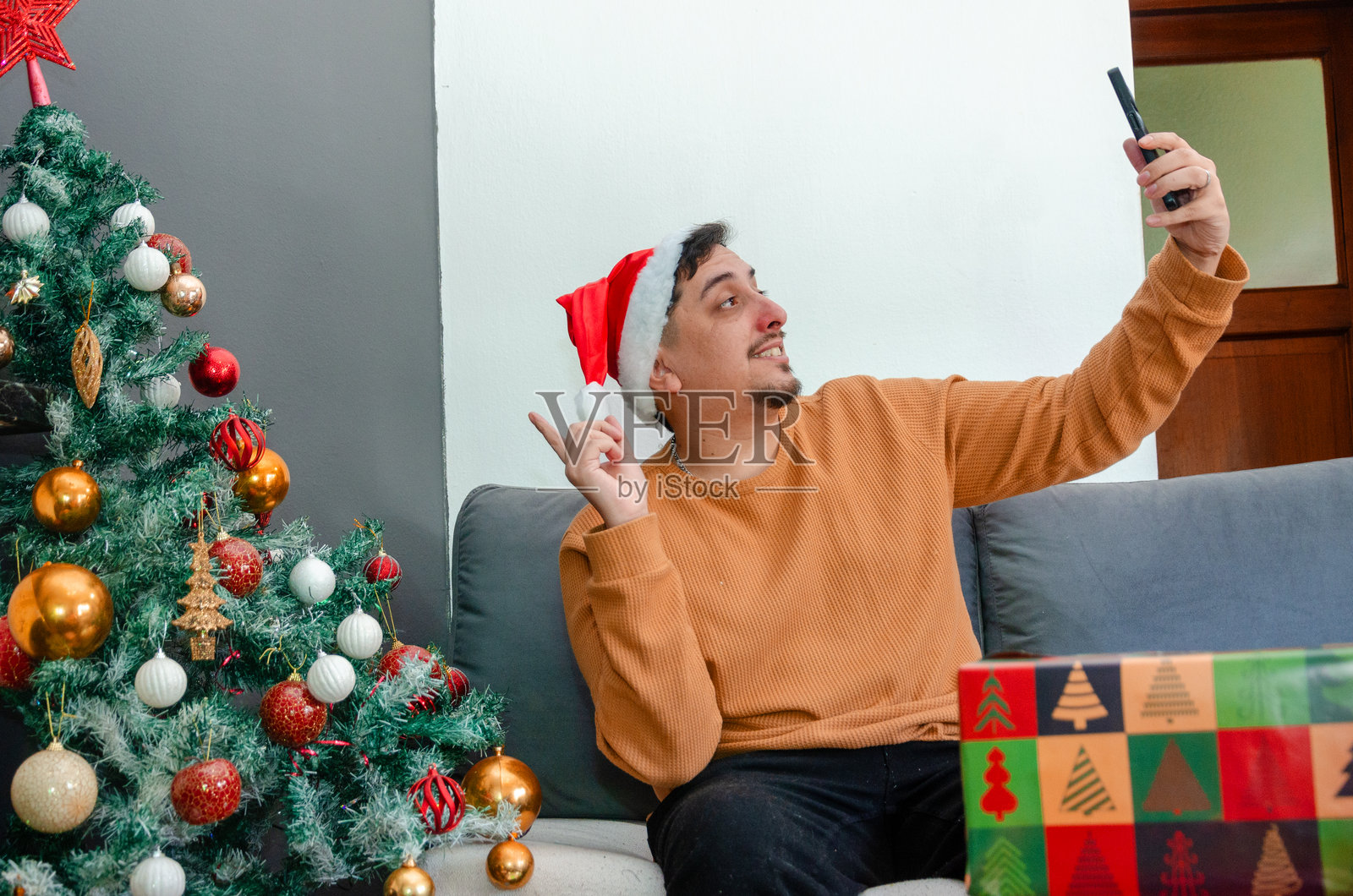 年轻男子戴着圣诞帽，用手机自拍，背景是圣诞树。照片摄影图片