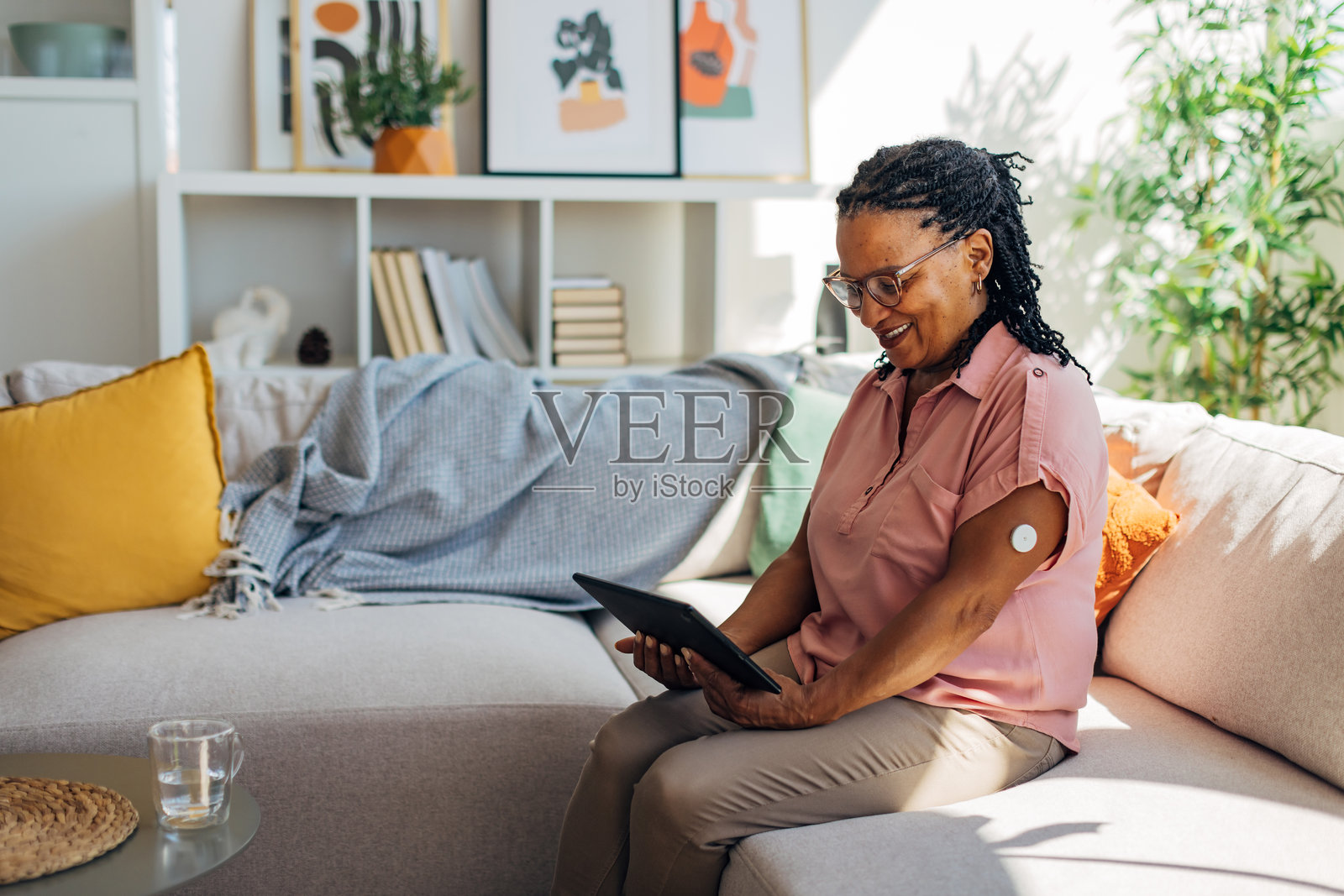 一位非裔美国女性在客厅放松，手臂上可见连续血糖监测设备，同时她正在浏览数字平板电脑。照片摄影图片