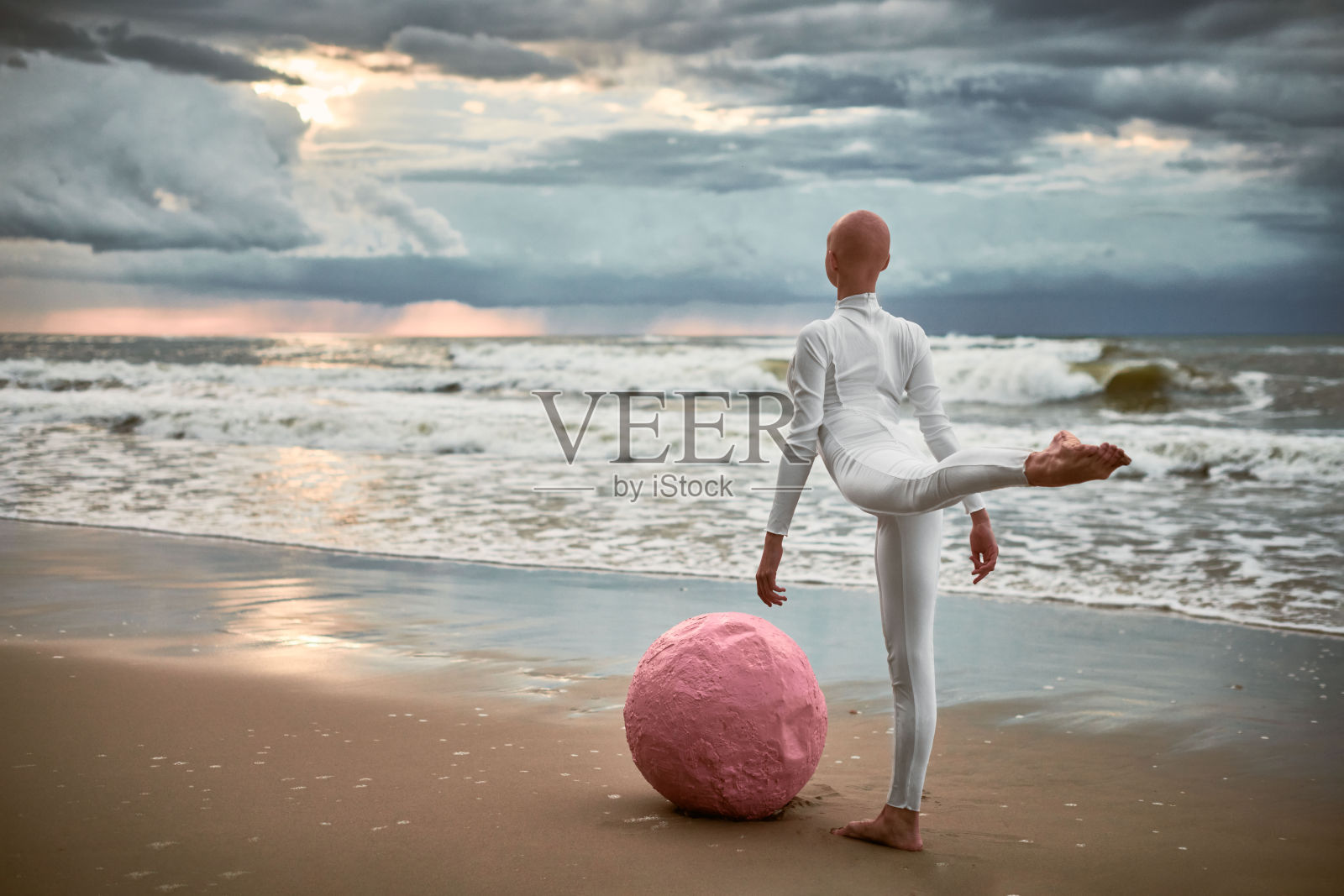 光头女孩在海滩上穿着白色未来主义套装，手持粉色球体跳舞。照片摄影图片