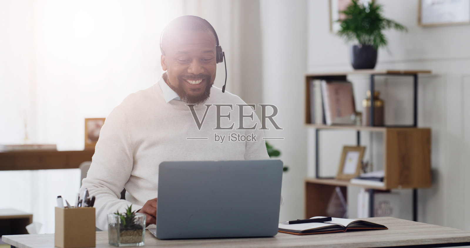 心理学家，佩戴耳机，在办公室通过笔记本电脑进行虚拟会议以提供建议，远程医疗或咨询。医生，成熟的黑人男性，愉快地在诊所进行视频通话，关注康复进展、治疗或后续跟进。照片摄影图片