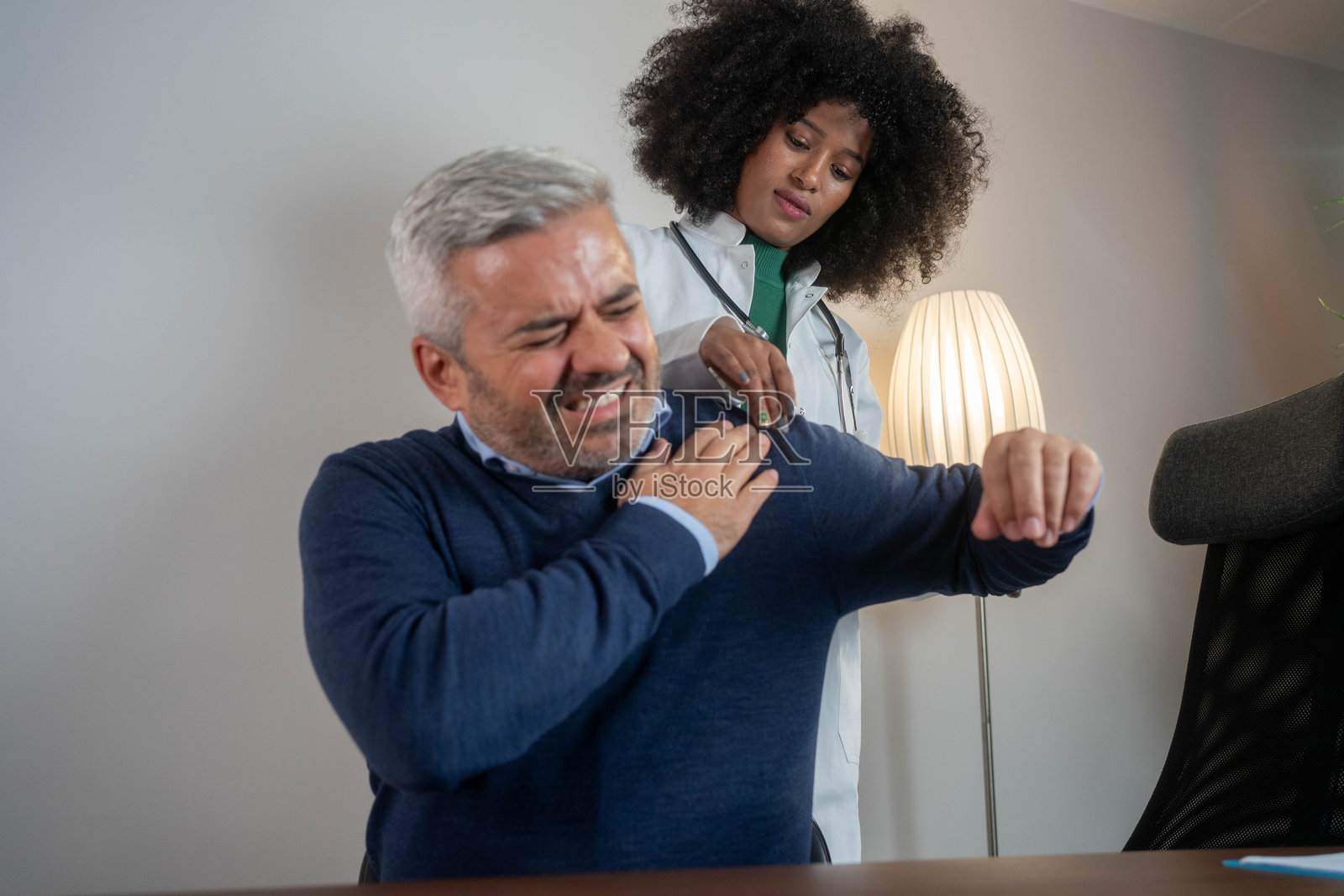 一名肩部疼痛的男性在医疗办公室咨询一位非洲女性医生，面露痛苦表情进行健康检查。照片摄影图片