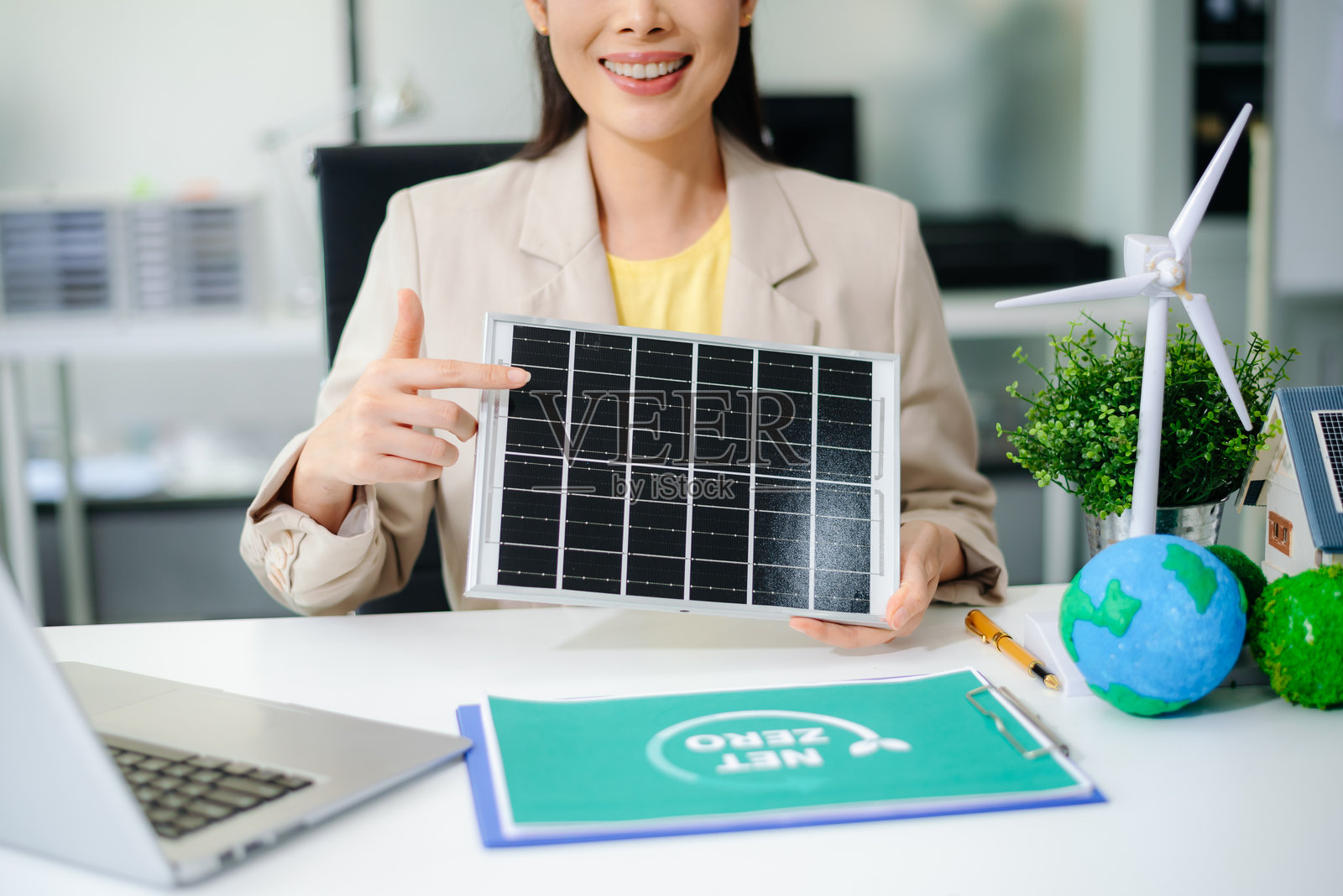 亚洲女性在生态办公室中手持太阳能电池板，周围有风力涡轮机、地球模型和净零文件夹。清洁能源、可再生技术和可持续商业概念。照片摄影图片