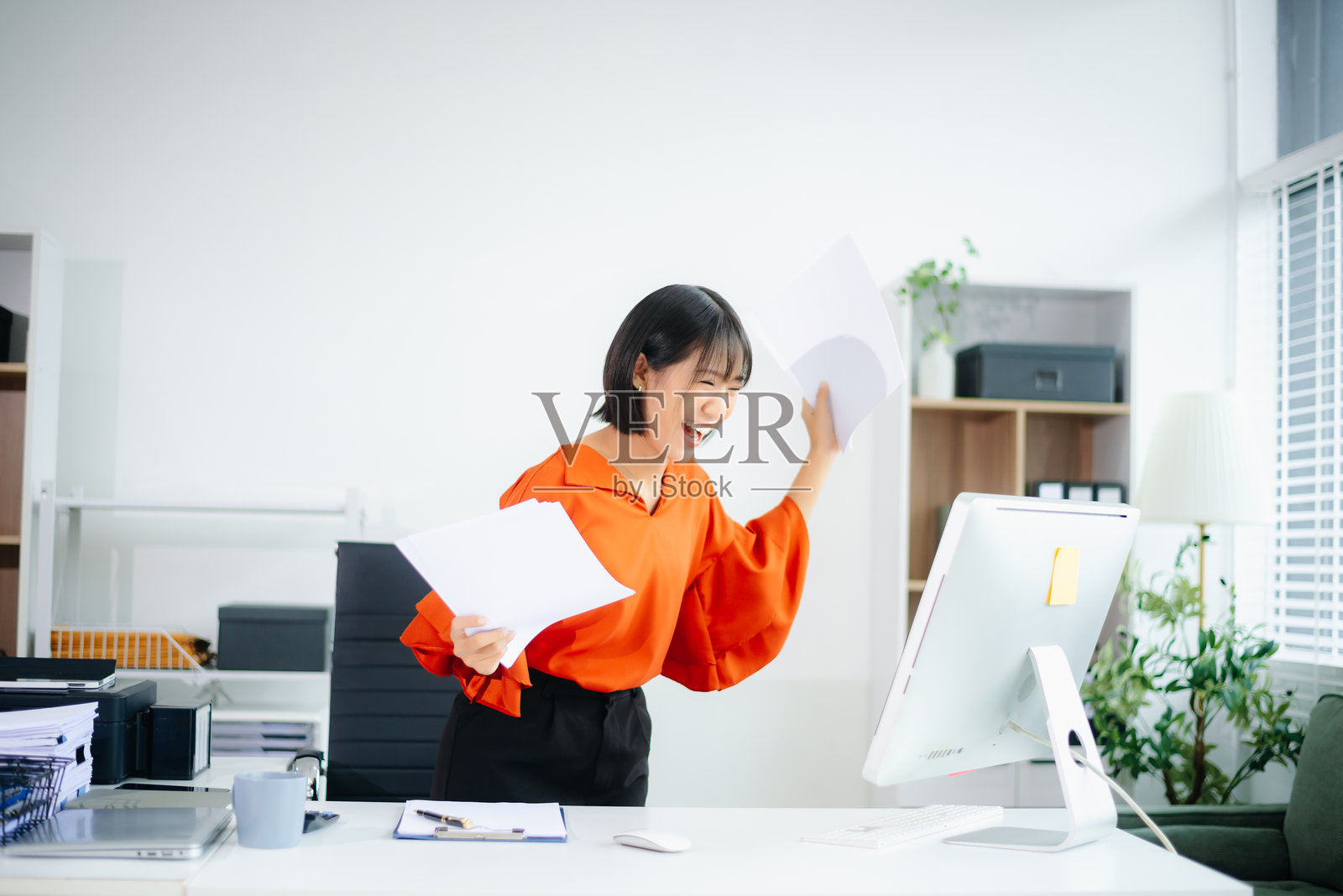 微笑的亚洲女性在明亮的办公室中举起文件庆祝成功，体现了商业胜利、快乐、激励和成就的概念。照片摄影图片