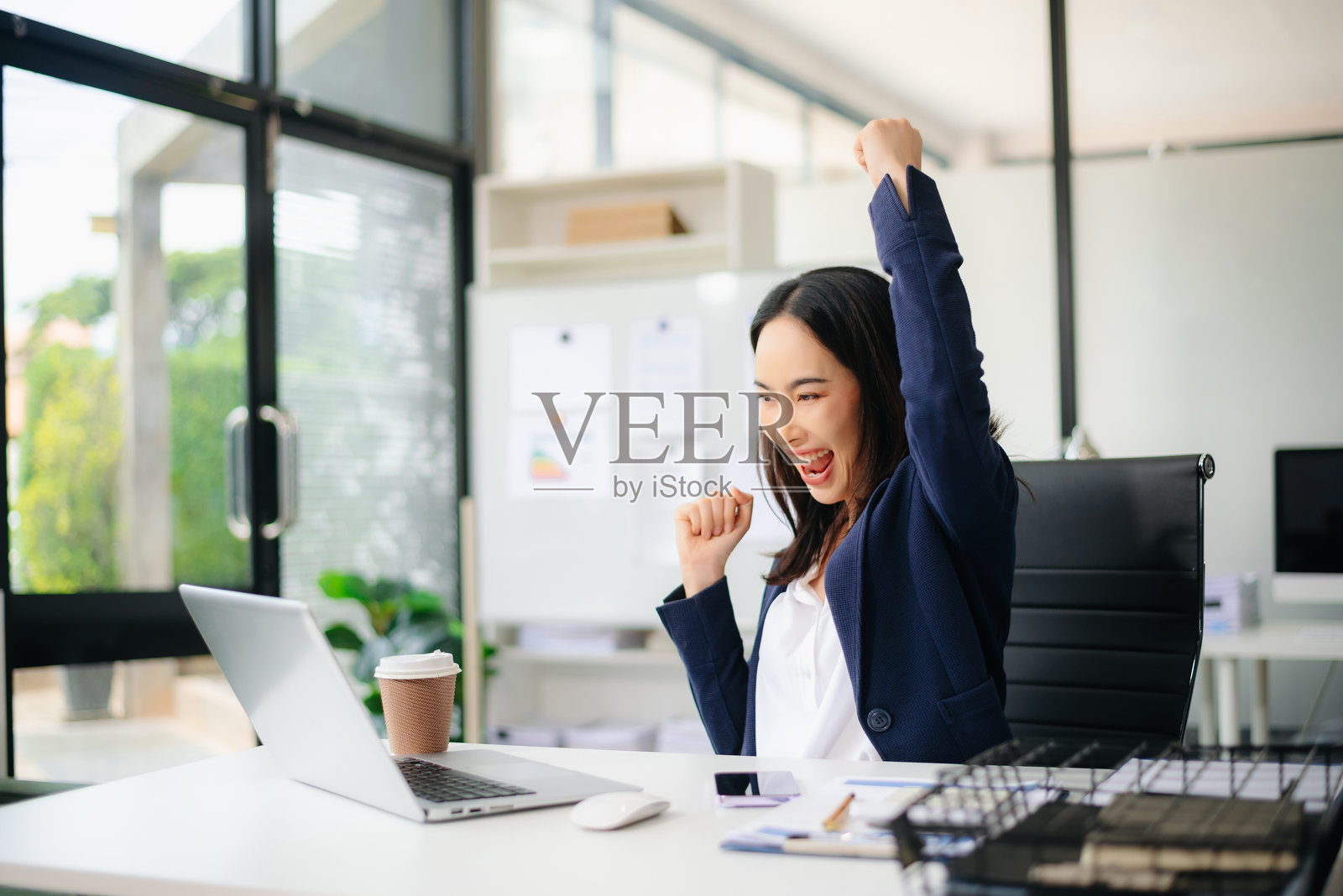 亚洲女商人在办公室桌前举起拳头庆祝成功，面前有一台笔记本电脑。照片摄影图片
