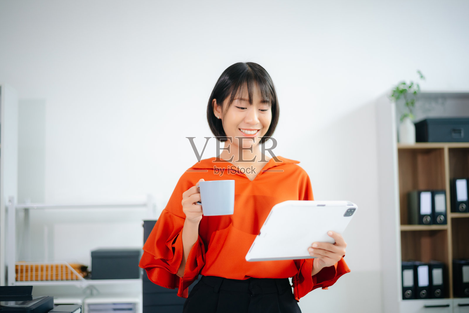 微笑的亚洲女性在明亮的办公室里查看平板电脑，同时手里拿着咖啡杯。远程工作和数字生活方式的概念。照片摄影图片