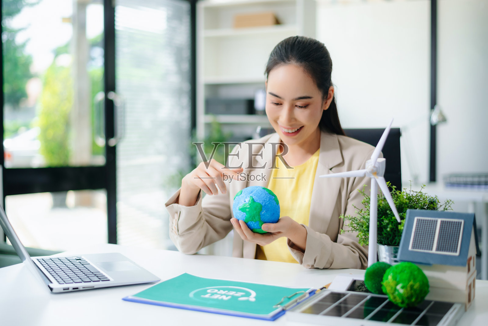 亚洲女性在使用笔记本电脑，手中拿着绿色星球模型，旁边有风力发电机和太阳能房屋。清洁能源的概念。照片摄影图片