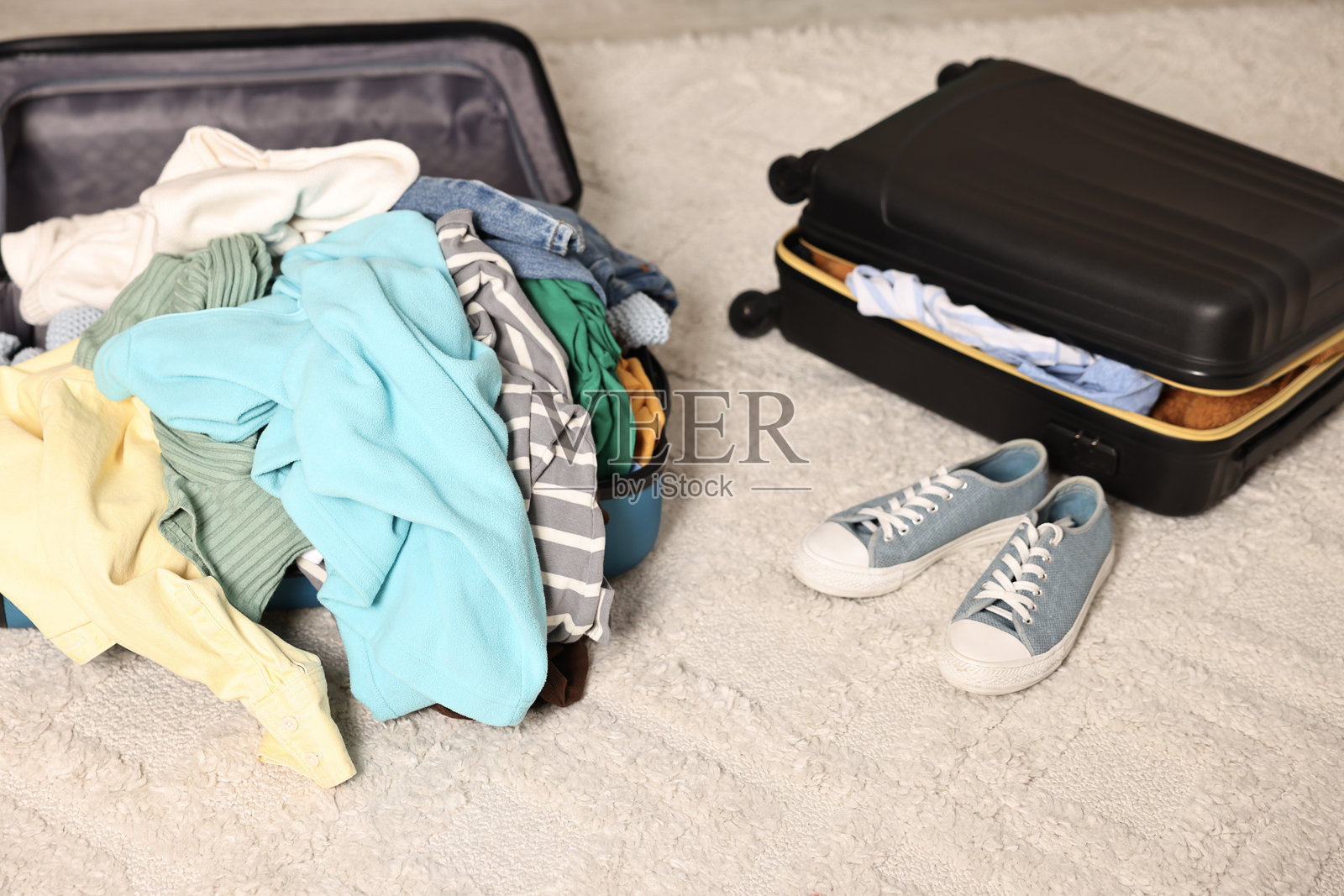 行李箱里堆满了不同的杂乱衣物，旁边还有一个行李箱和一双运动鞋，放在地毯上。照片摄影图片