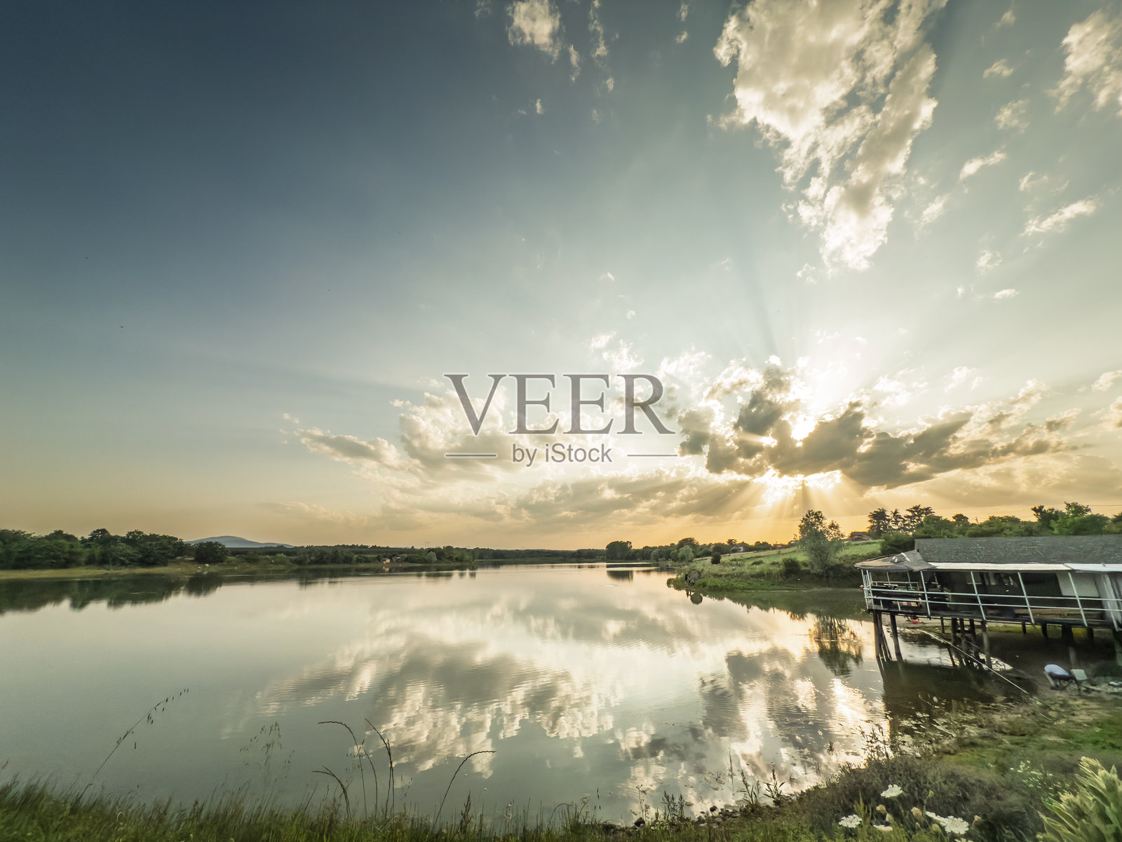 塞尔维亚姆拉登诺瓦茨附近的马尔科瓦茨湖上空的日落，阳光透过云层洒在平静的湖面上，湖边有一座小型高架建筑，草地岸边勾勒出宁静的乡村全景。照片摄影图片